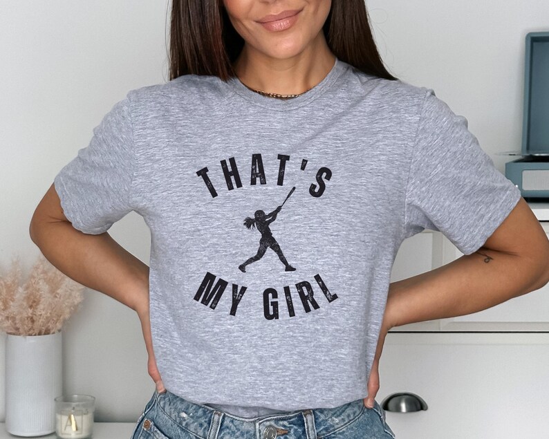 May include: Gray t-shirt with a black graphic of a girl playing baseball and the text "That's My Girl".