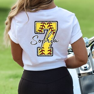 May include: White t-shirt with a yellow softball number 7 with red stitching and the name "Sophia" written in black cursive.