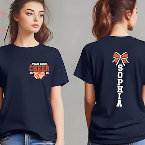 May include: A navy blue short-sleeve t-shirt featuring a front graphic with the text TEAM NAME CHEER 2025 and a pom-pom icon. The back of the shirt displays a large orange and white cheer bow graphic above the vertical white text SOPHIA.