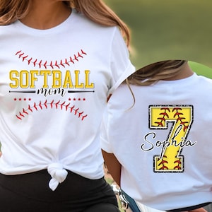 May include: Two white t-shirts with a softball theme. One shirt says "Softball Mom" with a red and white baseball stitch design. The other shirt has a large number 7 with a softball inside and the name "Sophia" underneath.