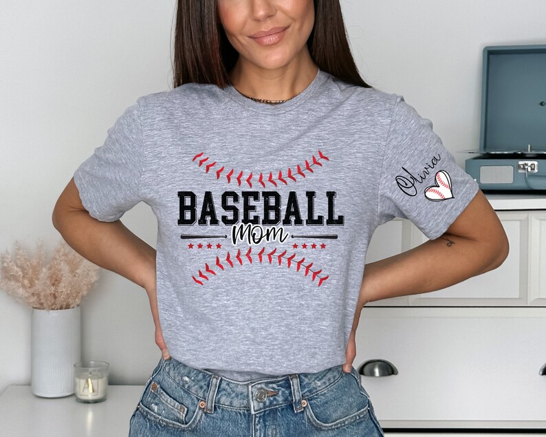 Peut inclure: T-shirt gris avec le texte "Baseball Mom" et un graphique de baseball. Le t-shirt a un c&oelig;ur rouge avec un graphique de baseball sur la manche et le nom "Olivia" &eacute;crit au-dessus.