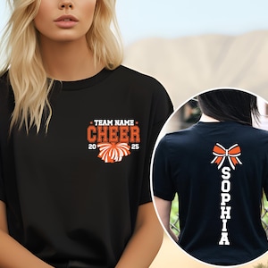 May include: A black t-shirt featuring a front graphic with the text TEAM NAME CHEER 2025 and an orange pom-pom. The back graphic displays a white and orange bow above the vertical text SOPHIA in white block letters on a black t-shirt background.