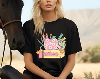 May include: A black t-shirt featuring a graphic print of a pink apple with white flowers, a yellow pencil with the name Alice written on it, crayons, a ruler, and a small bag. The design includes orange flowers and school supply illustrations.