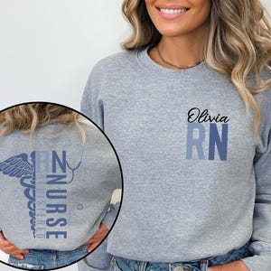 May include: A gray sweatshirt with a blue and white medical caduceus graphic on the back and the text "RN" with the name "Olivia" above it on the front.
