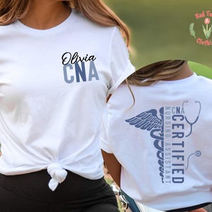 以下が含まれることがあります： 前面に「Olivia」と「CNA」とプリントされた白いTシャツ。Tシャツの背面には、医療用カドゥケウス、聴診器、および「CNA Certified Nursing Assistant」の文字が描かれています。