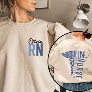 May include: A beige sweatshirt with the text "Olivia RN" printed on the front. The sweatshirt has a blue and white gradient design. The back of the sweatshirt features a stylized caduceus with the text "RN NURSE REGISTERED" printed vertically.