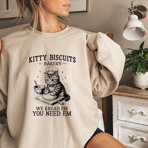 May include: A beige sweatshirt with a black graphic that reads "Kitty Biscuits Bakery" above an illustration of a cat wearing a chef's hat. The text "We Knead Em You Need Em" is below the cat illustration.