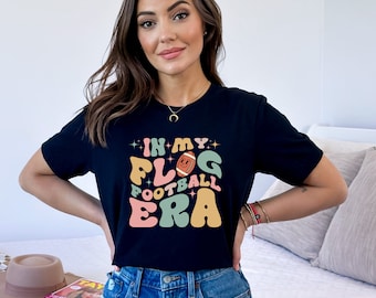 May include: A black t-shirt with a retro-style graphic that says "In My Football Era" with a cartoon football in the middle. The graphic is in shades of pink, yellow, green, and blue.