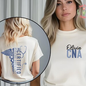 以下が含まれることがあります： クリーム色のTシャツ。黒い筆記体で「Olivia」と書かれ、その下に青い文字で「CNA」と書かれています。Tシャツの背面には、青いカドゥケウスのシンボルと「CNA CERTIFIED NURSING ASSISTANT」の文字、聴診器が描かれています。