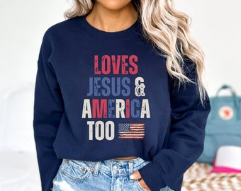 Może przedstawiać: Granatowa bluza z przetartym nadrukiem w kolorze białym, czerwonym i niebieskim, na którym widnieje napis "Loves Jesus & America Too" oraz mała amerykańska flaga.