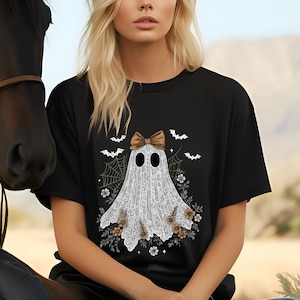 May include: Black t-shirt featuring a ghost design with a brown bow, spiderwebs, and bats. The ghost is white with a lace-like pattern. Floral accents in brown and white surround the ghost. The shirt is worn by a person.