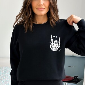 K&ouml;nnte beinhalten: Schwarzer Pullover mit einer wei&szlig;en Skeletthand, die die Rock'n'Roll-Handgeste macht. Die Hand ist von kleinen wei&szlig;en Sternen umgeben.