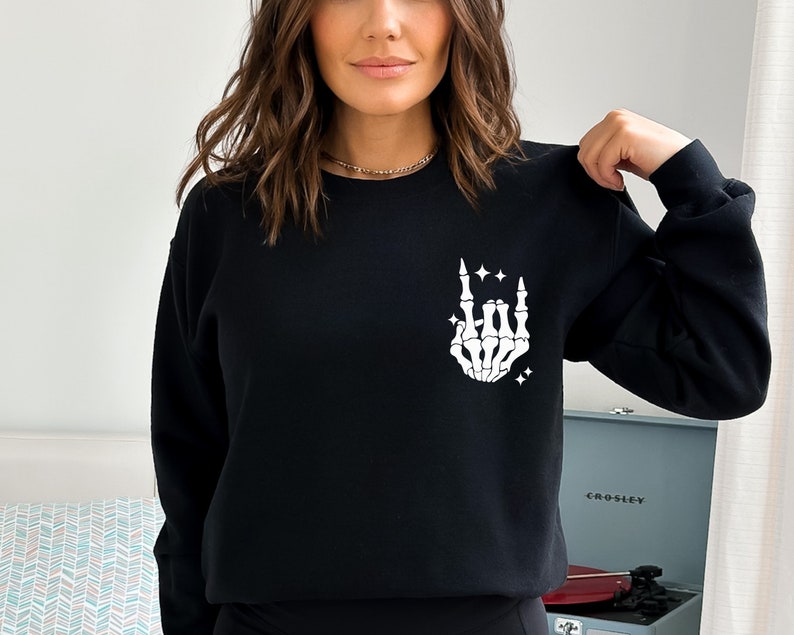 K&ouml;nnte beinhalten: Schwarzer Pullover mit einer wei&szlig;en Skeletthand, die die Rock'n'Roll-Handgeste macht. Die Hand ist von kleinen wei&szlig;en Sternen umgeben.