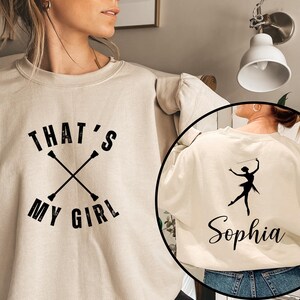 May include: Cream-colored sweatshirt with the text "THAT'S MY GIRL" in black, along with crossed batons. A circular inset shows the back of the sweatshirt with a black silhouette of a baton twirler and the name "Sophia".