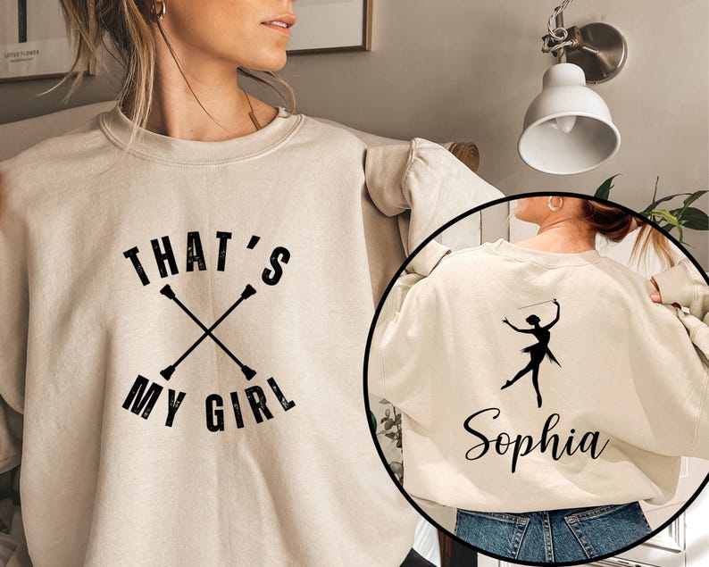 May include: Cream-colored sweatshirt with the text "THAT'S MY GIRL" in black, along with crossed batons. A circular inset shows the back of the sweatshirt with a black silhouette of a baton twirler and the name "Sophia".