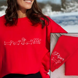 K&ouml;nnte beinhalten: Roter Pullover mit einem wei&szlig;en Umriss einer Reihe von Dackeln. Der Pullover hat den Namen "Piper" auf dem &Auml;rmel gestickt.