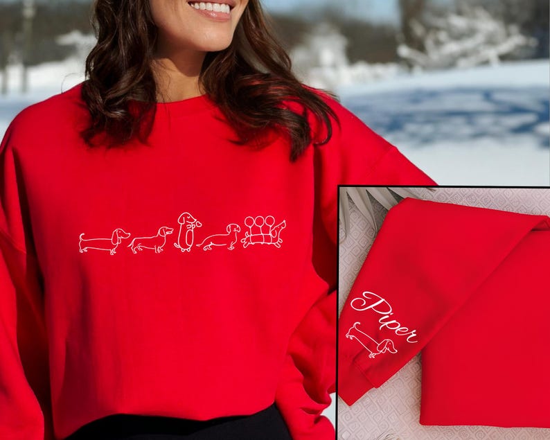 K&ouml;nnte beinhalten: Roter Pullover mit einem wei&szlig;en Umriss einer Reihe von Dackeln. Der Pullover hat den Namen "Piper" auf dem &Auml;rmel gestickt.