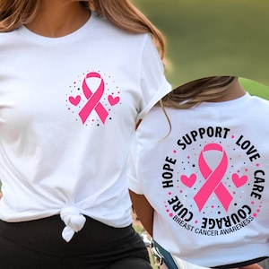 Könnte beinhalten: Weißes T-Shirt mit einem rosa Band und zwei Herzen auf der Vorderseite. Die Rückseite des Shirts hat ein rosa Band mit den Worten "HOPE, SUPPORT, LOVE, CARE, COURAGE, CURE, BREAST CANCER AWARENESS" drumherum.