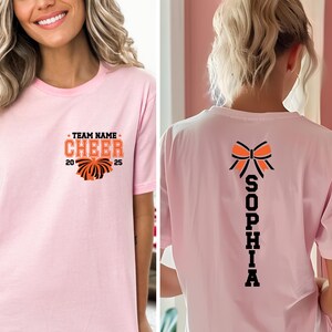 May include: A light pink t-shirt featuring a front graphic with the text TEAM NAME CHEER 2025 and a pom-pom illustration. The back of the shirt displays a large orange and black bow graphic above the vertical text SOPHIA in bold black letters.