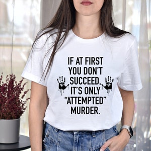 May include: White t-shirt with black text that reads "If at first you don't succeed it's only "attempted" murder."  Two black handprints with dripping blood are on either side of the text.