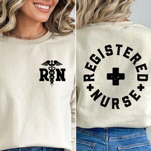 May include: A beige sweatshirt with a black medical symbol and the letters "RN" on the front. The back of the sweatshirt has a black circle with the words "Registered Nurse" in a vintage style font.