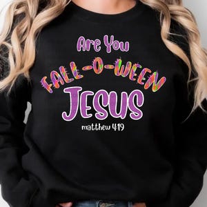 Puede incluir: Sudadera negra con el texto "Are You FALL-O-WEEN JESUS" en letras moradas y coloridas. El texto "matthew 4:19" está debajo de la palabra "JESUS". Un diseño religioso con temática de Halloween.