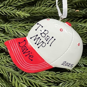 May include: A baseball cap ornament with a white crown and a red brim. The text "T-Ball MVP Davie 2025" is written on the cap. The ornament is hanging from a white ribbon against a backdrop of green pine needles.