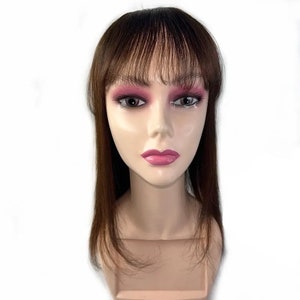 May include: A mannequin head with a brown wig featuring a fringe and long, straight hair. The mannequin has a fair complexion, with pink eyeshadow and lipstick. The wig appears to be a natural shade of brown.
