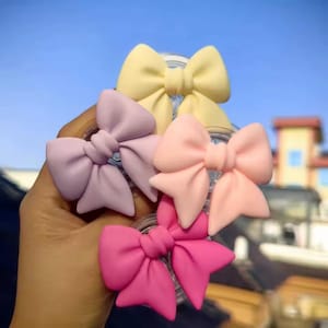 May include: A collection of colorful hair bows in shades of yellow, lavender, peach, and hot pink. The bows are made of a soft material and are arranged in a hand against a blue sky background.