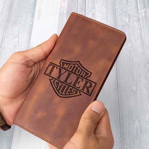 May include: A brown leather wallet with the text "MOTOR TYLER CYCLES" engraved on the front. The wallet is rectangular and held in two hands. The leather has a distressed appearance.