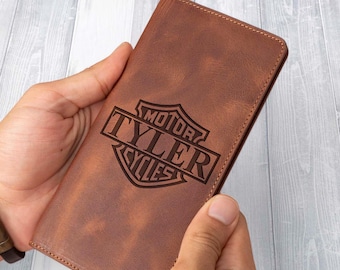 May include: A brown leather wallet with the text "MOTOR TYLER CYCLES" engraved on the front. The wallet is rectangular and held in two hands. The leather has a distressed appearance.