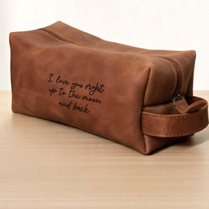 Puede incluir: Un neceser de cuero marrón con cierre de cremallera y asa. La bolsa tiene el texto "I love you right up to the moon and back." Un recipiente de "got2b GENTLEMAN TEXTURE CLAY" está al lado de la bolsa.