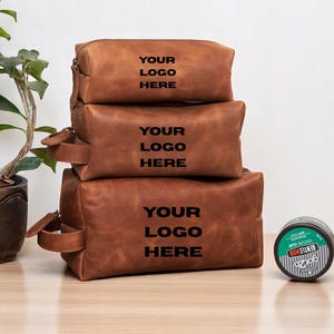 Puede incluir: Tres neceseres de cuero marrón de diferentes tamaños están apilados sobre una superficie de madera. Cada bolsa tiene cierre de cremallera y asa. El texto "YOUR LOGO HERE" está impreso en cada bolsa. Una lata verde y negra y una botella marrón están junto a las bolsas.