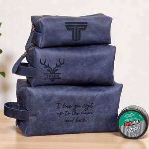May include: Three blue leather wash bags stacked, with hair clay and beard balm. The top bag has a "T" logo, the middle bag has a deer logo with "TYLER", and the bottom bag has the text "I love you right up to the moon and back." The products are on a wooden surface.