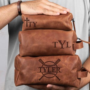 Puede incluir: Tres neceseres de cuero marrón apilados. La bolsa superior tiene las iniciales "TY" grabadas. La bolsa del medio tiene "TYL" grabado. La bolsa inferior tiene un diseño de béisbol con el nombre "TYLER" grabado.