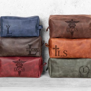 Puede incluir: Una pila de neceseres de cuero en varios colores, incluyendo azul, marrón y rojo. Cada bolsa presenta texto y símbolos personalizados, como una corona, un símbolo médico, una cruz y una corona de laurel, junto con el nombre "Lucas" o iniciales.
