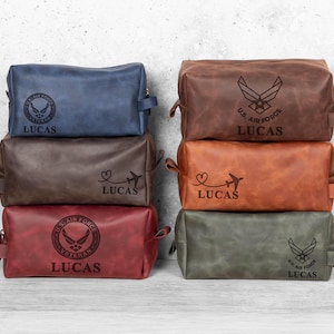 May include: A stack of six leather toiletry bags in various colors: navy blue, brown, burgundy, dark brown, orange-brown, and olive green. Each bag has a zipper closure, a handle, and the name "LUCAS" engraved. Some bags feature U.S. Air Force emblems.