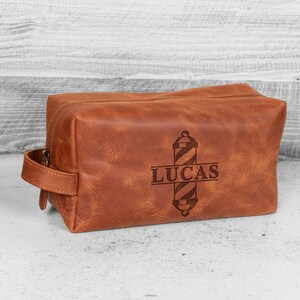 Puede incluir: Un neceser de cuero marr&oacute;n con asa y cierre de cremallera. La bolsa presenta un dise&ntilde;o de poste de barbero y el nombre "LUCAS" grabado en la parte delantera. La bolsa es rectangular.