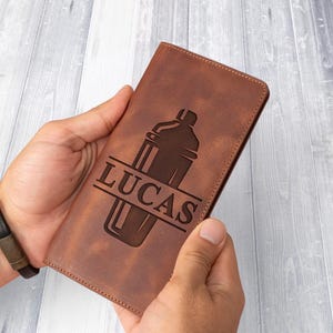 May include: A brown leather wallet with a cocktail shaker design and the name "LUCAS" engraved on the front. The wallet is held in two hands, showcasing its rectangular shape and textured surface.