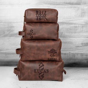 Puede incluir: Cuatro neceseres de cuero marr&oacute;n apilados. Cada bolsa tiene un dise&ntilde;o diferente con tem&aacute;tica de barber&iacute;a y el nombre "LUCAS" grabado. Las bolsas son rectangulares con cierre de cremallera y un asa lateral.