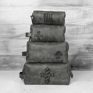 Puede incluir: Cuatro neceseres de cuero gris apilados. Cada bolsa tiene un dise&ntilde;o diferente con tem&aacute;tica de barber&iacute;a y el nombre "LUCAS" grabado. Las bolsas son de diferentes tama&ntilde;os y tienen cierre de cremallera.