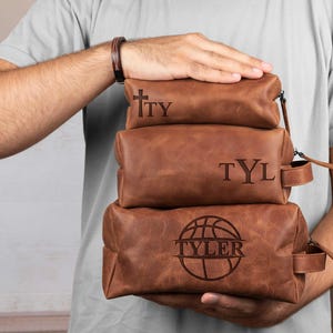 Puede incluir: Tres neceseres de cuero marrón apilados. La bolsa superior tiene las iniciales "TY" grabadas, la bolsa del medio "TYL", y la bolsa inferior presenta un gráfico de baloncesto y el nombre "TYLER".