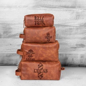 Puede incluir: Cuatro neceseres de cuero marr&oacute;n apilados con cremalleras y asas. Cada bolsa tiene un dise&ntilde;o diferente con tem&aacute;tica de barber&iacute;a y el nombre "LUCAS" grabado. Est&aacute;n colocados sobre un fondo de madera blanca y gris.