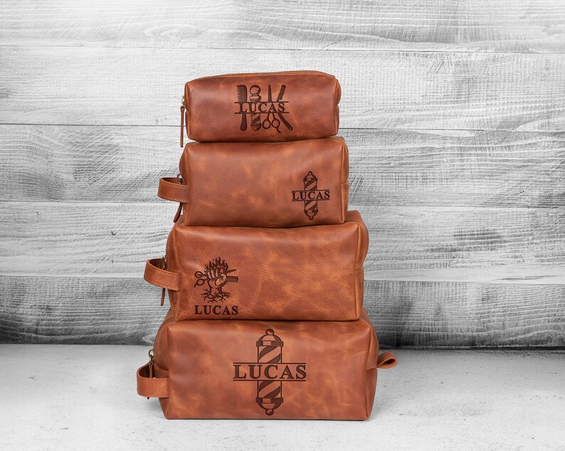 Puede incluir: Cuatro neceseres de cuero marr&oacute;n apilados con cremalleras y asas. Cada bolsa tiene un dise&ntilde;o diferente con tem&aacute;tica de barber&iacute;a y el nombre "LUCAS" grabado. Est&aacute;n colocados sobre un fondo de madera blanca y gris.