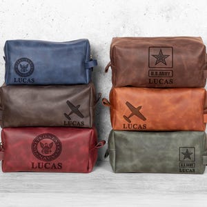 Puede incluir: Seis neceseres de cuero en varios colores: azul, marrón, rojo, beige y verde. Cada bolsa tiene cierre de cremallera, asa y nombre personalizado. Algunas bolsas presentan emblemas militares y un avión.
