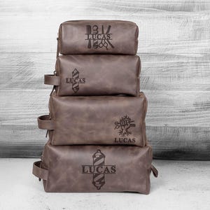 Puede incluir: Cuatro neceseres de cuero marr&oacute;n apilados con texto personalizado y dise&ntilde;os tem&aacute;ticos de barber&iacute;a. Cada bolsa tiene un dise&ntilde;o diferente, incluyendo tijeras, un poste de barbero y el nombre "LUCAS". Las bolsas tienen un asa lateral.