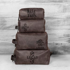Puede incluir: Cuatro neceseres de cuero marr&oacute;n apilados con cremalleras y asas negras. Cada bolsa tiene un gr&aacute;fico diferente con tem&aacute;tica de barber&iacute;a y el nombre "LUCAS" grabado. Sobre un fondo de madera blanca.