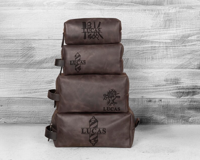 Puede incluir: Cuatro neceseres de cuero marr&oacute;n apilados con cremalleras y asas negras. Cada bolsa tiene un gr&aacute;fico diferente con tem&aacute;tica de barber&iacute;a y el nombre "LUCAS" grabado. Sobre un fondo de madera blanca.