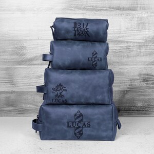 Puede incluir: Cuatro neceseres de cuero azul apilados, con dise&ntilde;os grabados en negro y el nombre "LUCAS". Cada bolsa tiene un dise&ntilde;o diferente, incluyendo un poste de barbero, un &aacute;rbol y un emblema vintage. Las bolsas son de diferentes tama&ntilde;os y tienen un asa lateral.