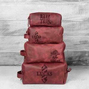 Puede incluir: Una pila de cuatro neceseres de cuero burdeos. Cada bolsa tiene cierre de cremallera y asa. Las bolsas est&aacute;n grabadas con dise&ntilde;os de barber&iacute;a y el nombre "LUCAS".